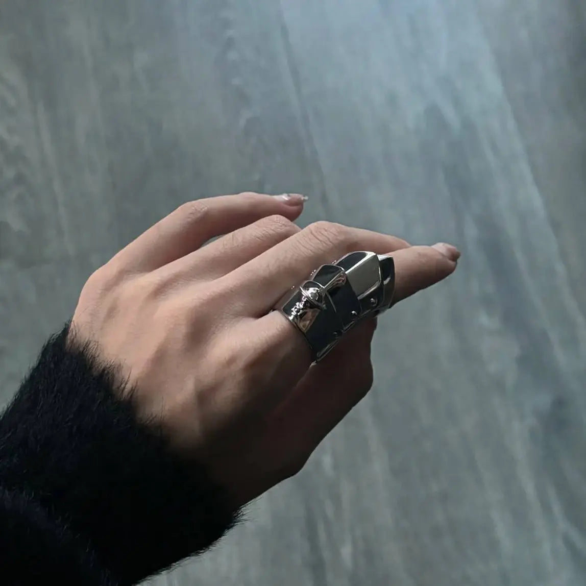 A close-up of a hand wearing a chunky silver-toned geometric ring with a textured black band, set against a gray wood-look floor background.