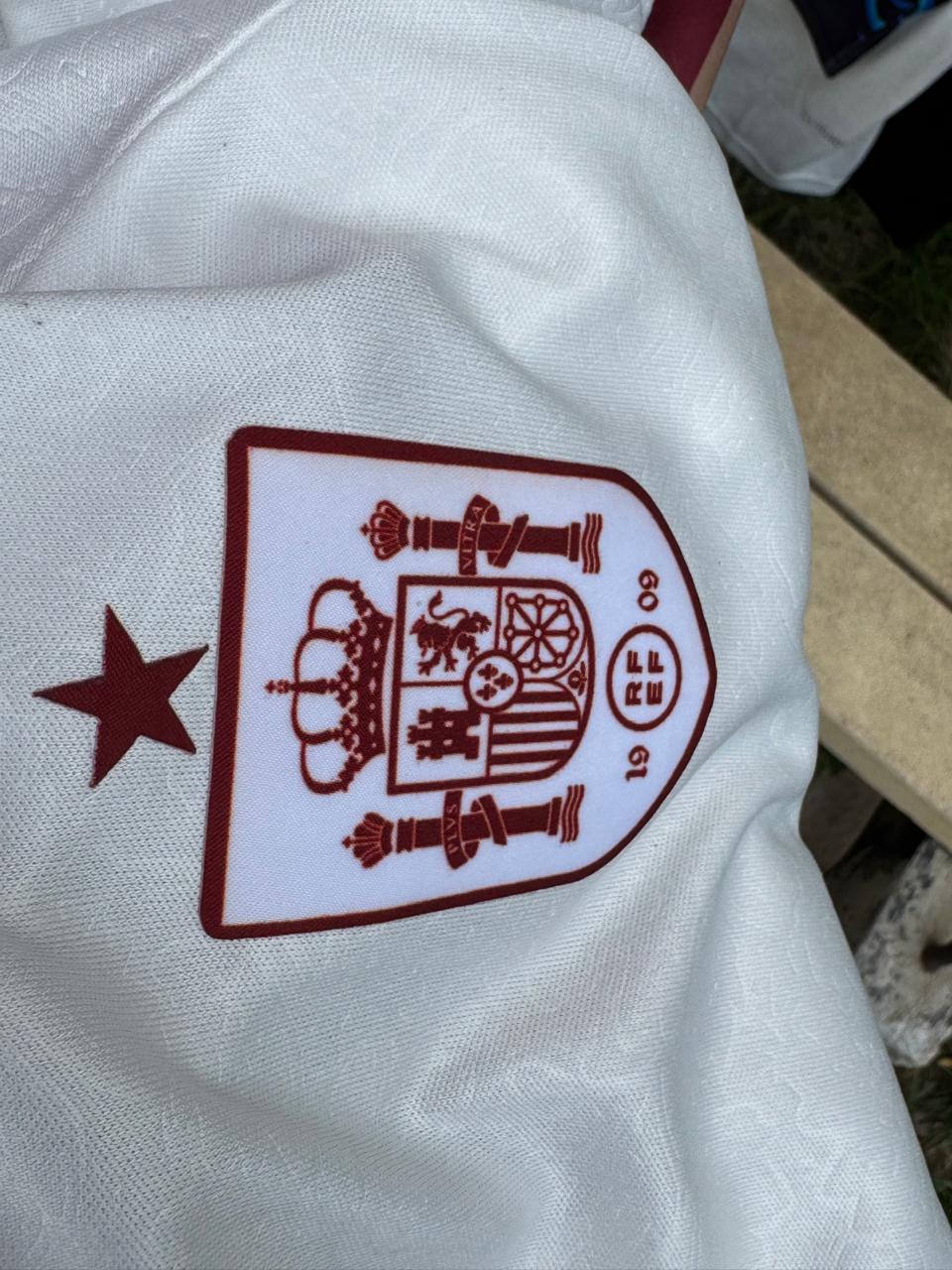 Close-up of a white textured fabric item featuring the Spain national team crest in maroon, with a maroon star on the left.