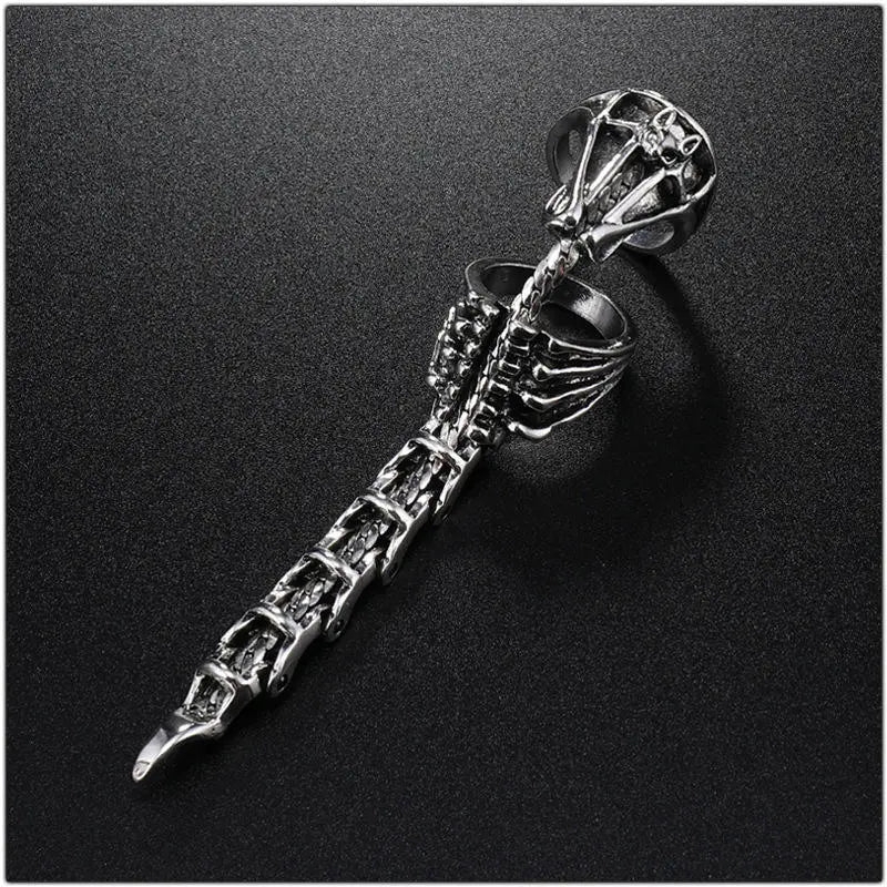 A silver-toned gothic ring featuring a detailed skeleton hand holding a sword, displayed diagonally on a dark textured background.